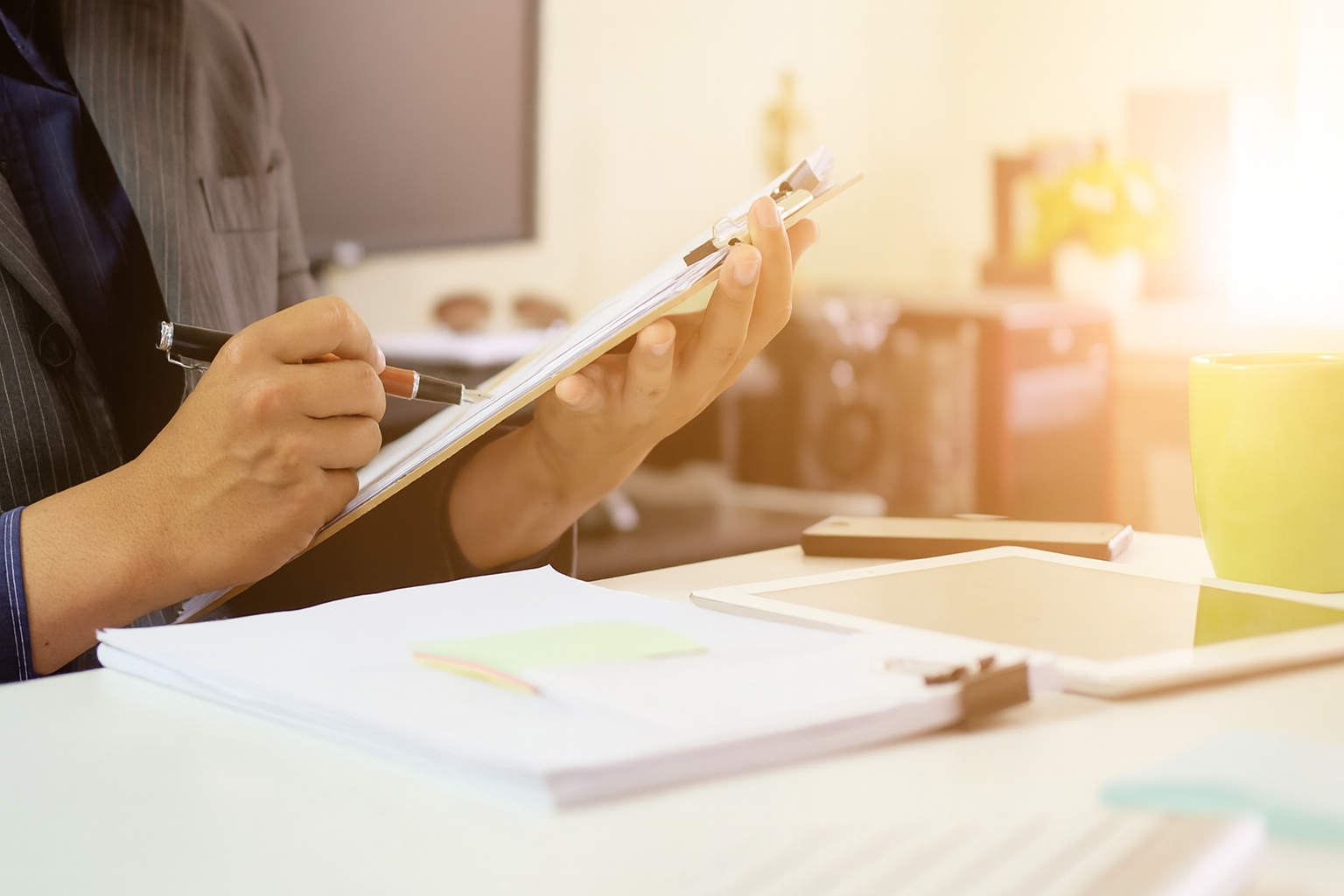 A person in a business suit sits at a desk, reviewing documents on a clipboard with a pen in hand. The workspace includes papers, a tablet, a smartphone, and a green coffee mug, suggesting a focused office environment.