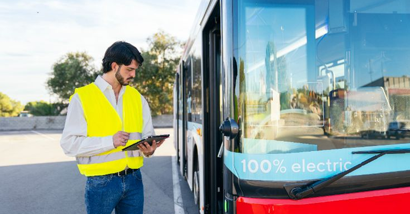 A safety‑vested worker stands beside an electric bus while reviewing information on a tablet.