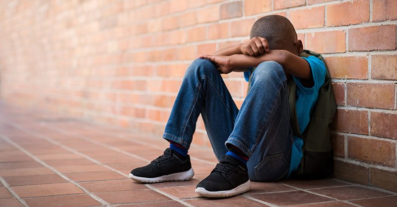 A sad boy sitting against a wall with his head resting on his arms
