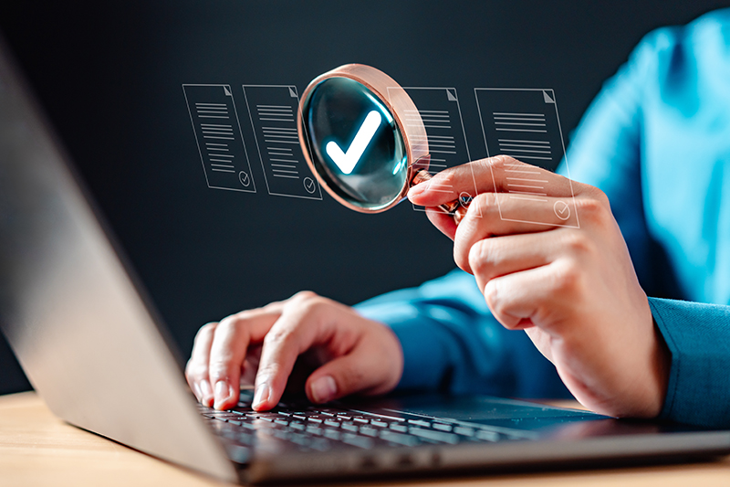 Document, information, paper, text, examining, find, inspect, investigation, management, research. A person is using laptop with magnifying glass, on a document that is being checked.