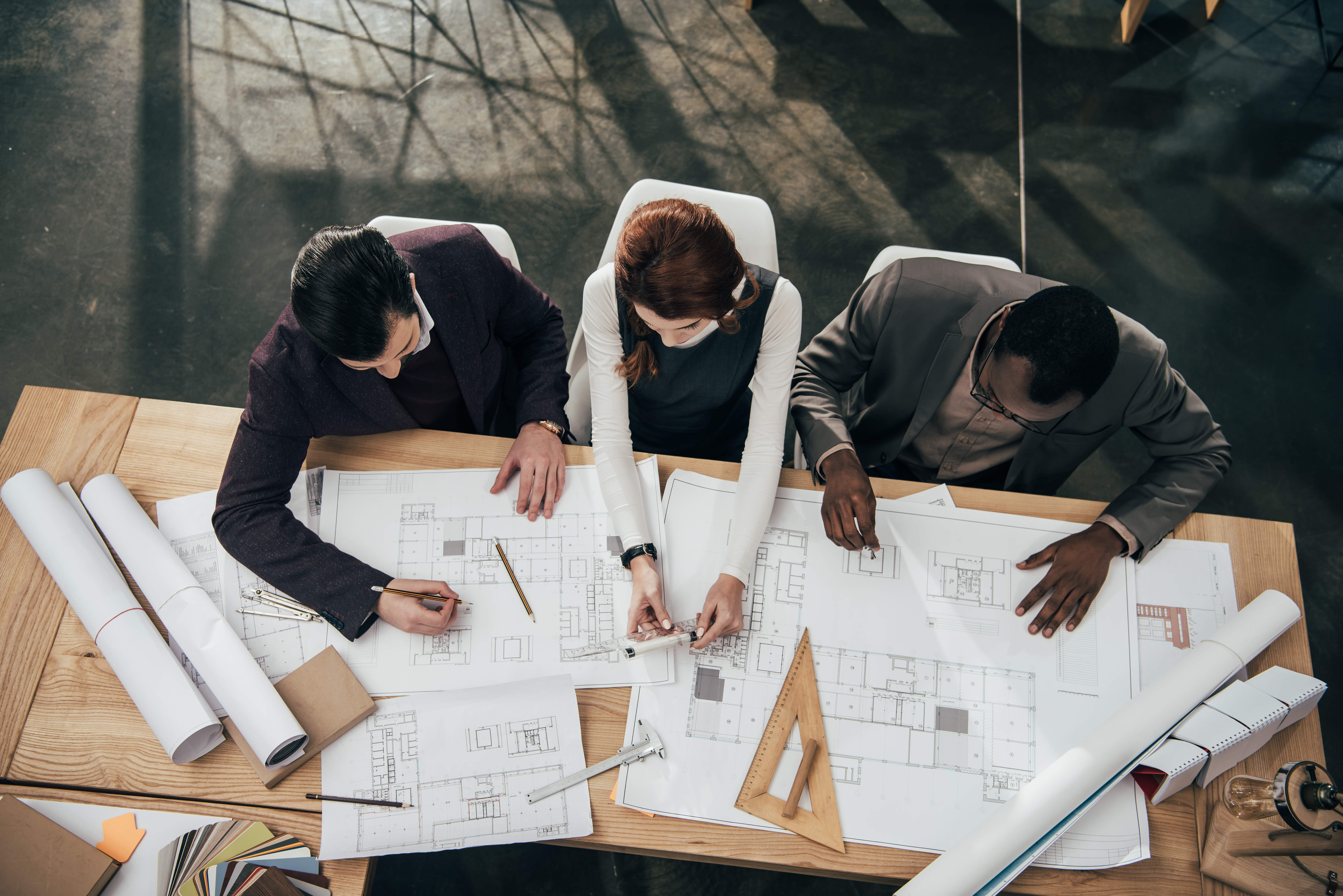 Three people sit at a wooden table reviewing architectural blueprints, surrounded by drafting tools like a triangular ruler. They are engaged in discussion, pointing at various sections of the plans.