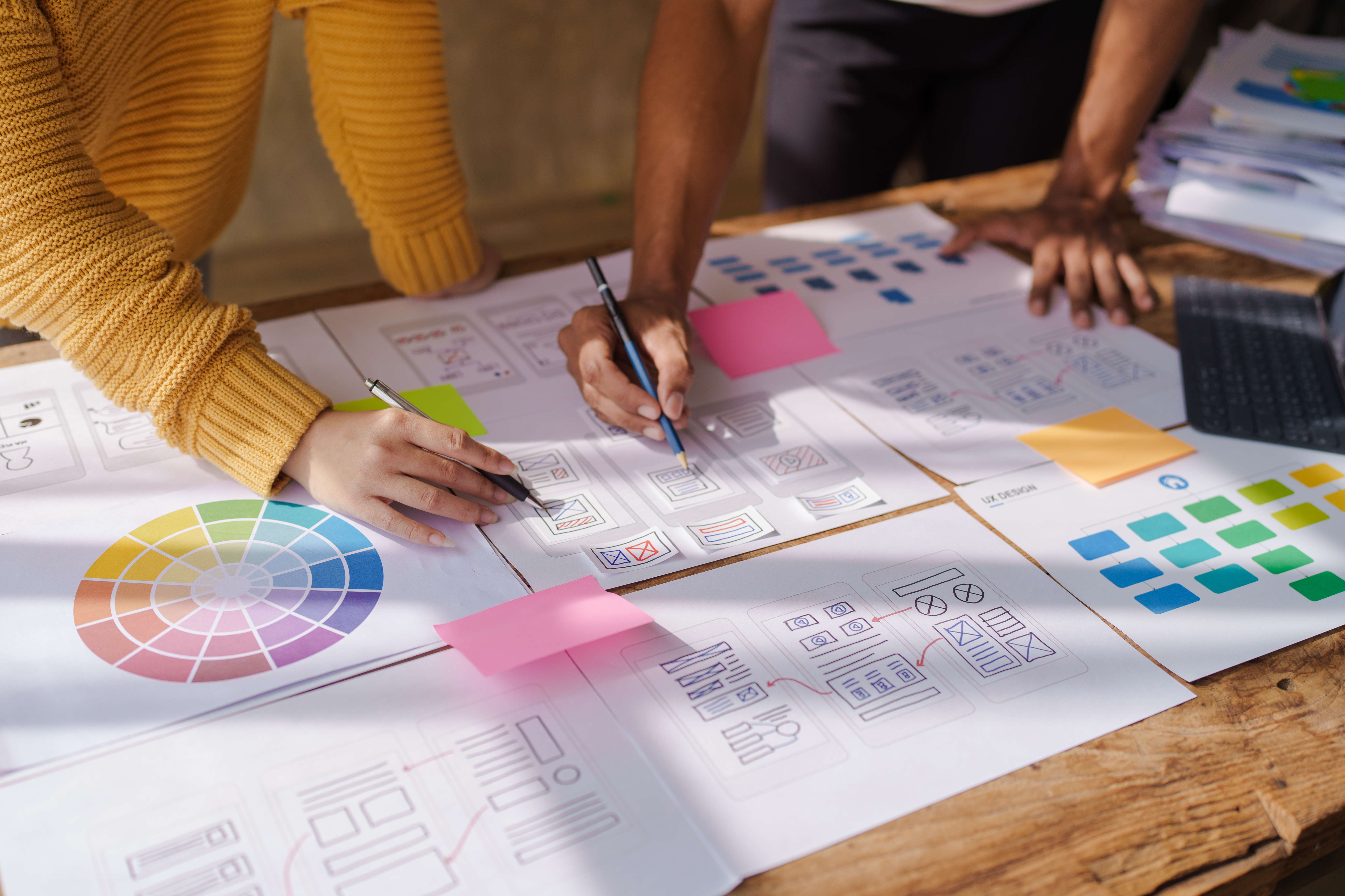 Two people collaborate at a wooden table filled with design sketches, color palettes, sticky notes, a laptop, and papers. One wears a yellow sweater and holds a pen; only the other’s arm is visible.