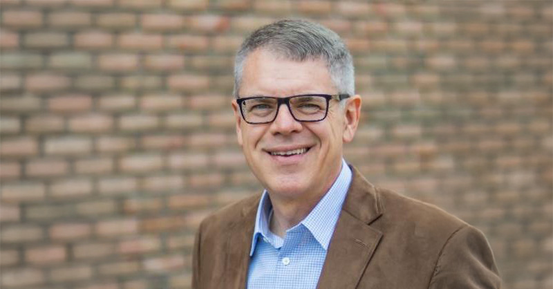 A person wearing a light blue checkered button-up shirt under a brown suede blazer, standing outdoors in front of a brick wall background.